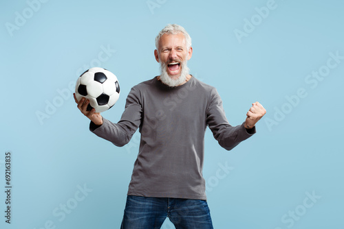 Bearded attractive mature man holding soccer ball cheering, standing isolated on blue background © Maria Vitkovska