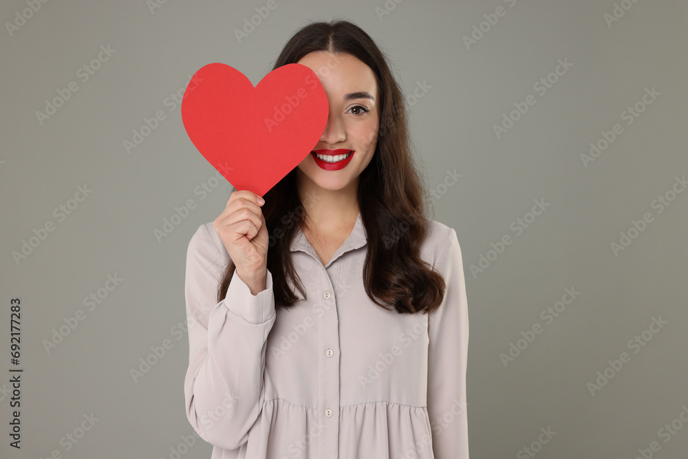 Beautiful young woman covering her eye with paper heart on grey background Beautiful young woman covering her eye with paper heart on grey background