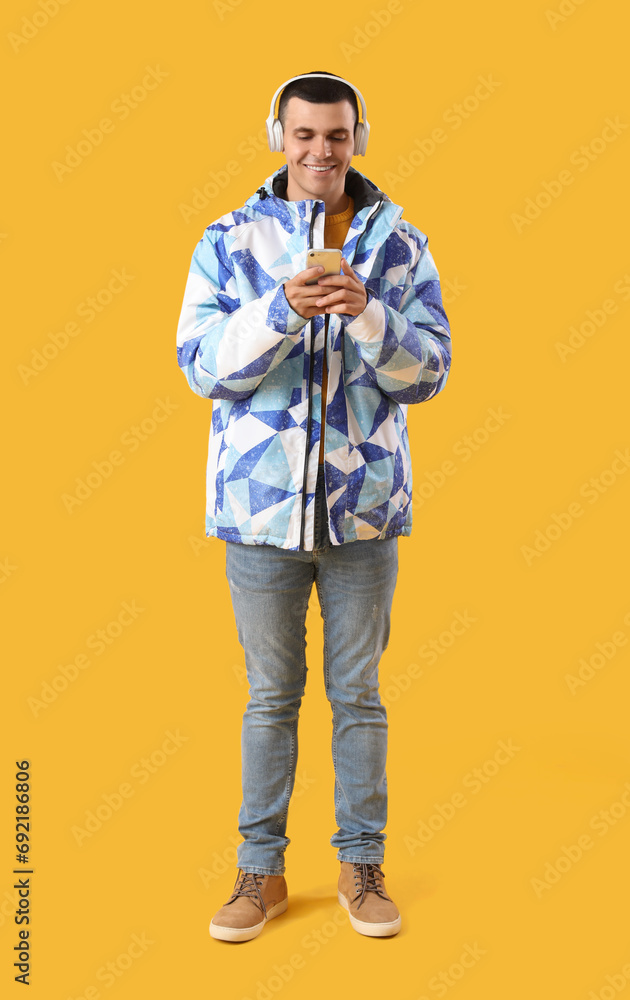 Young man in warm jacket with headphones using mobile phone on yellow background Young man in warm jacket with headphones using mobile phone on yellow background