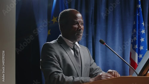 Senior African American politician in formal suit holding speech by podium with mic against EU and US flags during press conference photo