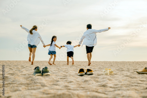 Back view of family parents with children fun holding hands together running to beach in holiday, dad, mom and kids take off shoes run on sand beach, daughter turned face around, Happy family day