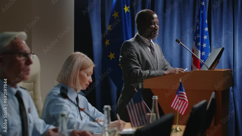 Mature Black political leader in formal suit standing by podium with mic against EU and US flags and giving speech during military committee conference