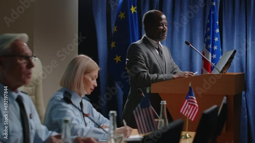 Mature Black political leader in formal suit standing by podium with mic against EU and US flags and giving speech during military committee conference photo