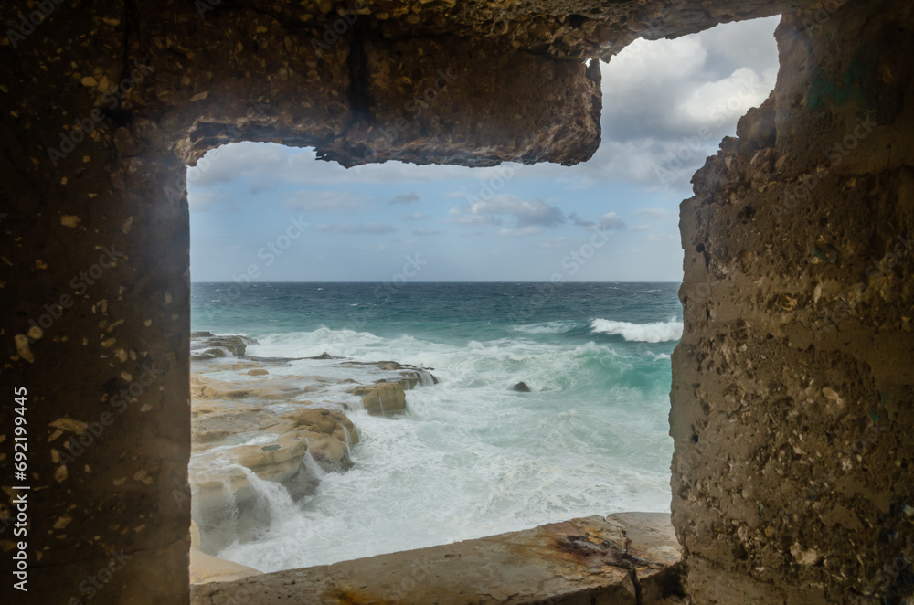 Rocky shore against a cloudy sky and waves in the sea in Malta. Photo taken through a window.