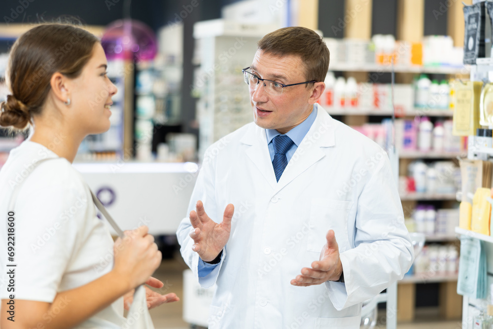 Obraz premium Positive young female and middle-aged male pharmacists talking to each-other in chemist's shop with large assortment