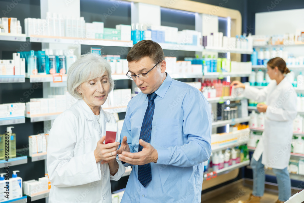 Polite mature female pharmacist consulting middle-aged man costumer about care product in box in chemist's shop