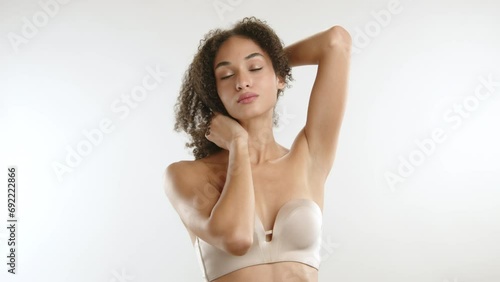 Beautiful confident lady with african curls posing on white background in non wired strapless bra of smooth fabric tossing brown hair, shaking head. High quality 4k footage