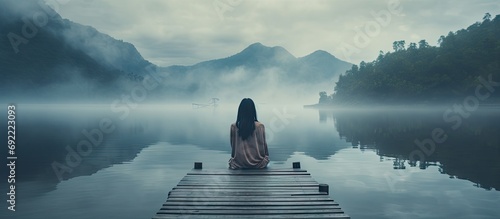 Back view of fashioned young woman sitting on wooden dock looking at view on a misty morning Female hipster with brown hat relaxes on the edge of jetty admiring foggy lake Wonderful nature geta photo