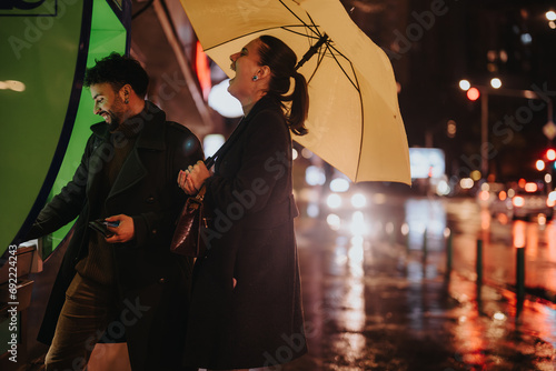 A couple using an ATM on a rainy day. The man withdraws money while holding an umbrella, while the woman stands beside him. They are stylishly dressed for business. photo