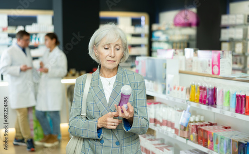 Near self-service window in pharmacy, senior female customer chooses lubricant gel, carefully reads information for consumer on packaging of product