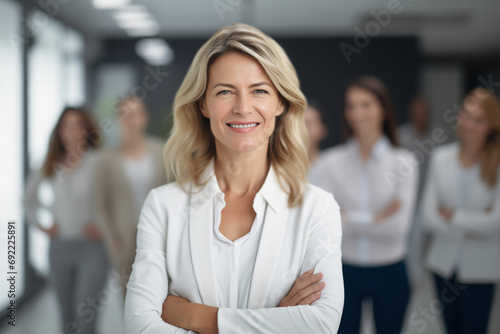business woman wearing white jacket suit, in the office, her work team appears in the background out of focus. smiling woman. boss, businesswoman, leader