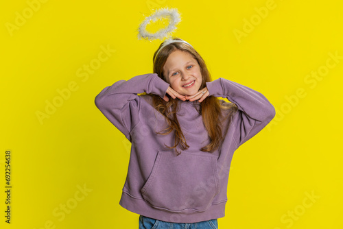 Portrait of smiling shy angelic young preteen child girl kid with angel halo nimb over head flirting, looking at camera, positive emotions, celebrating holiday. Children isolated on yellow background