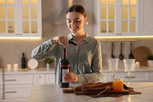 Romantic dinner. Woman opening wine bottle with corkscrew at table in kitchen photo