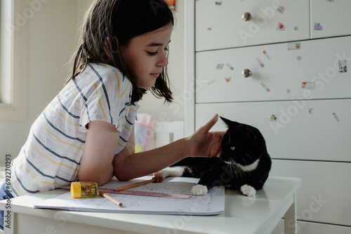 Young preteen girl in her bedroom remote home schooling, distracted patting her newly adopted kitten lying on her desk during covid-19 lockdown, Victoria, Australia; April 2020 photo