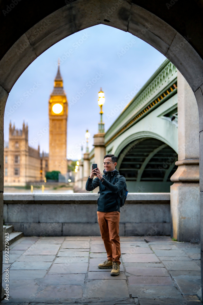 Full length of young Latin man tourist in casual clothes standing alone against ancient cathedral in London near tower bridge and Big Ben against clock tower and taking photos while sightseeing