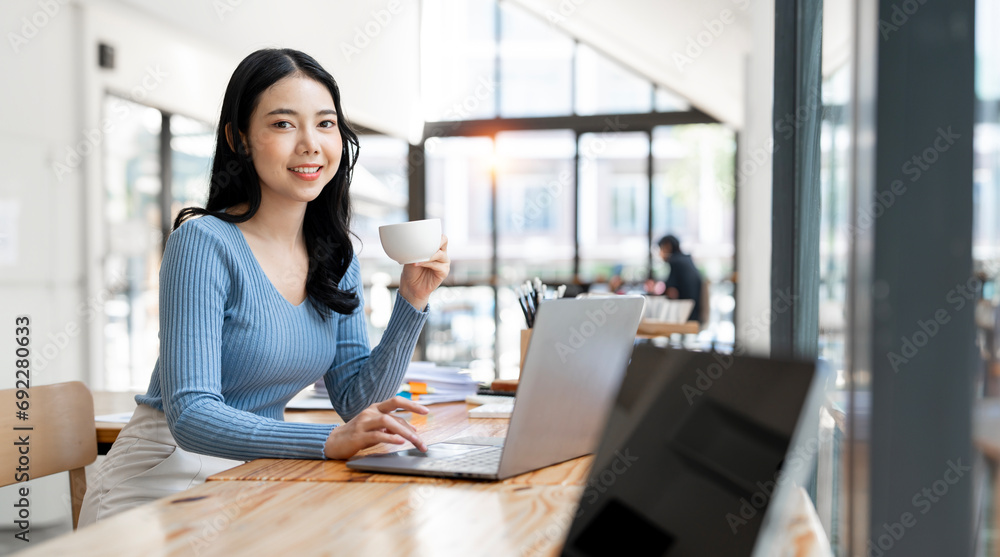 Smiling woman holding coffee cup and using laptop while sitting at co-workspace. Smiling woman holding coffee cup and using laptop while sitting at co-workspace.
