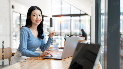 Smiling woman holding coffee cup and using laptop while sitting at co-workspace.