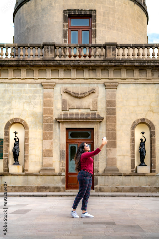 Happy young smiling Latin woman tourist in casual clothes and sunglasses taking selfie with smartphone while standing against blurred historic Chapultepec Castle in Mexico City during trip