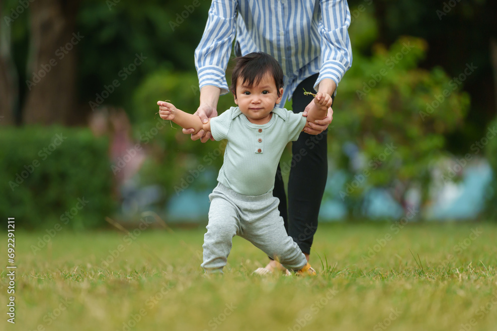 infant baby learn walking first step on grass field with mother holding hand helping in park