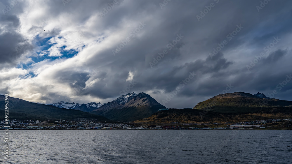A picturesque snow-capped mountain range of the Andes against a cloudy sky. The town houses of Ushuaia are visible at the foot of the mountains, on the banks of the Beagle Canal. Argentina