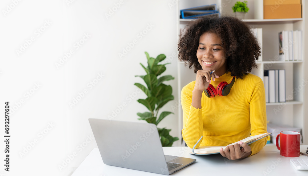 © ArLawKa - African American girl in headphones watching video lesson on computer in living room at home, happy in headphones having online class using laptop