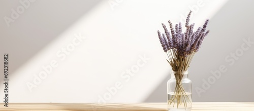 White dried lavender in clear glass vase on glossy black table with wooden trim. Placed against white wall with gentle shading, casting shadow on right side. photo