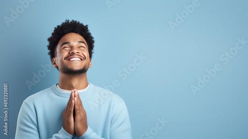 A man who has achieved success is praying and making wishes with his hands crossed, gazing up with a pleading expression and standing against a blue backdrop. photo
