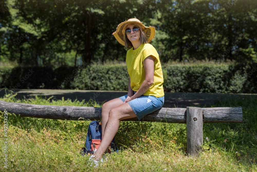A short rest for a walk in the summer park. The woman is blonde, wearing a yellow T-shirt and a straw hat.