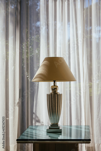 Elegant white antique table lamp with fabric lampshade on the glass table near the window. Lamp silhouette on the curtain in sunny day. Classic decoration for home or hotel architecture