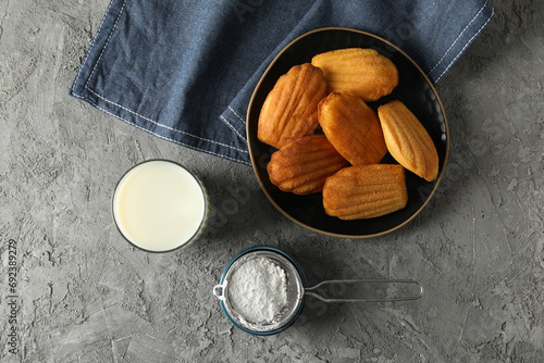 Madeleine cakes in bowl, glass of milk, flour and towel on gray background, top view photo