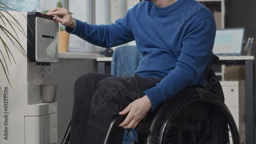 Tilt up shot of young Caucasian wheelchaired man pouring fresh still water from water dispenser into cup while during workday in modern office photo