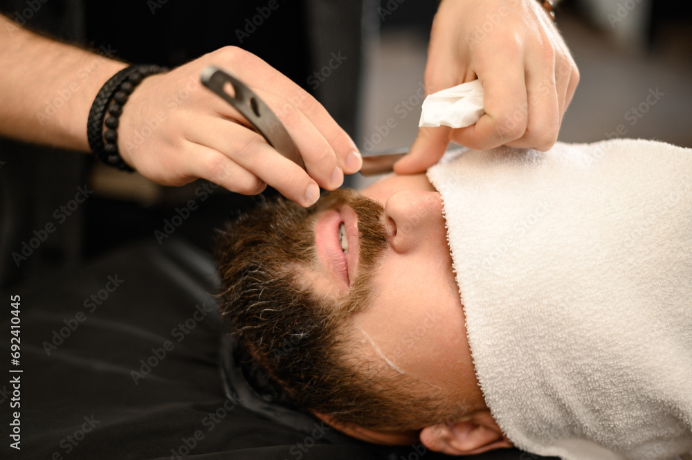 A barber shaves the cheek of a bearded customer with dangerous razor. Shaving the contour of the beard for the correct shape. A barber shaves the cheek of a bearded customer with dangerous razor. Shaving the contour of the beard for the correct shape.