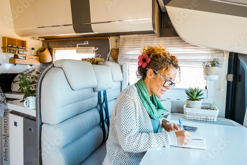 One woman sitting inside modern motorhome alternative tiny house and write travel notes on the table. People living vanlife on the road life and vacation. Traveler female indoor leisure van activity photo