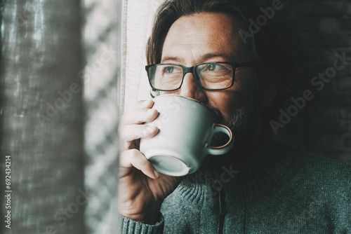 One man with thoughtful and serene expression on face drinking coffee or tea alone at home looking outside the window. Mature male people alone. Healthy mindful lifestyle concept. Wearing glasses photo