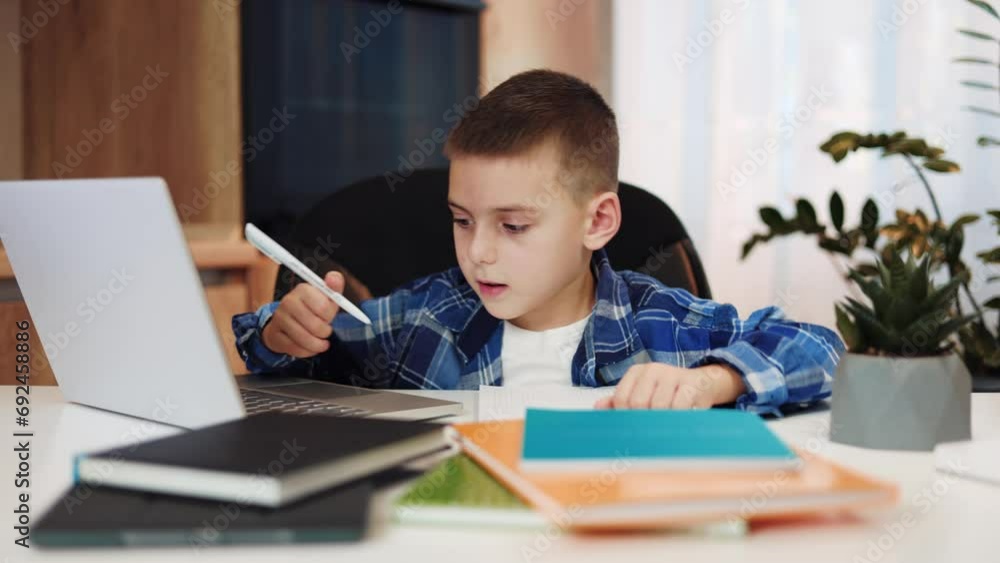 Attentive caucasian male child typing on portable laptop and writing something in personal notebook. Enthusiastic preschooler sitting at table with books and equipment for distance education at home. Attentive caucasian male child typing on portable laptop and writing something in personal notebook. Enthusiastic preschooler sitting at table with books and equipment for distance education at home.
