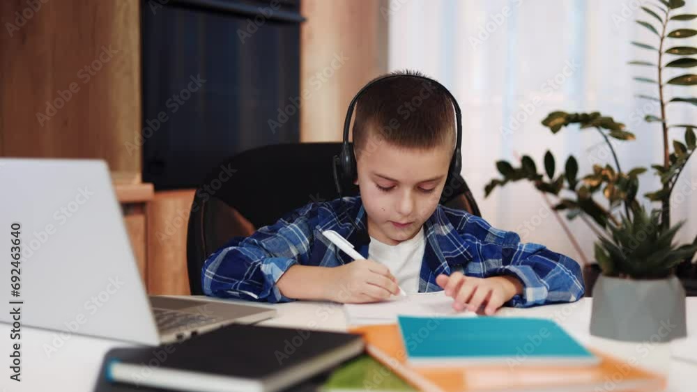 Elementary student in casual clothes using modern headset connected to laptop while homeschooling alone. Smart pupil taking notes and showing thumb up during video call during distance learning. Elementary student in casual clothes using modern headset connected to laptop while homeschooling alone. Smart pupil taking notes and showing thumb up during video call during distance learning.