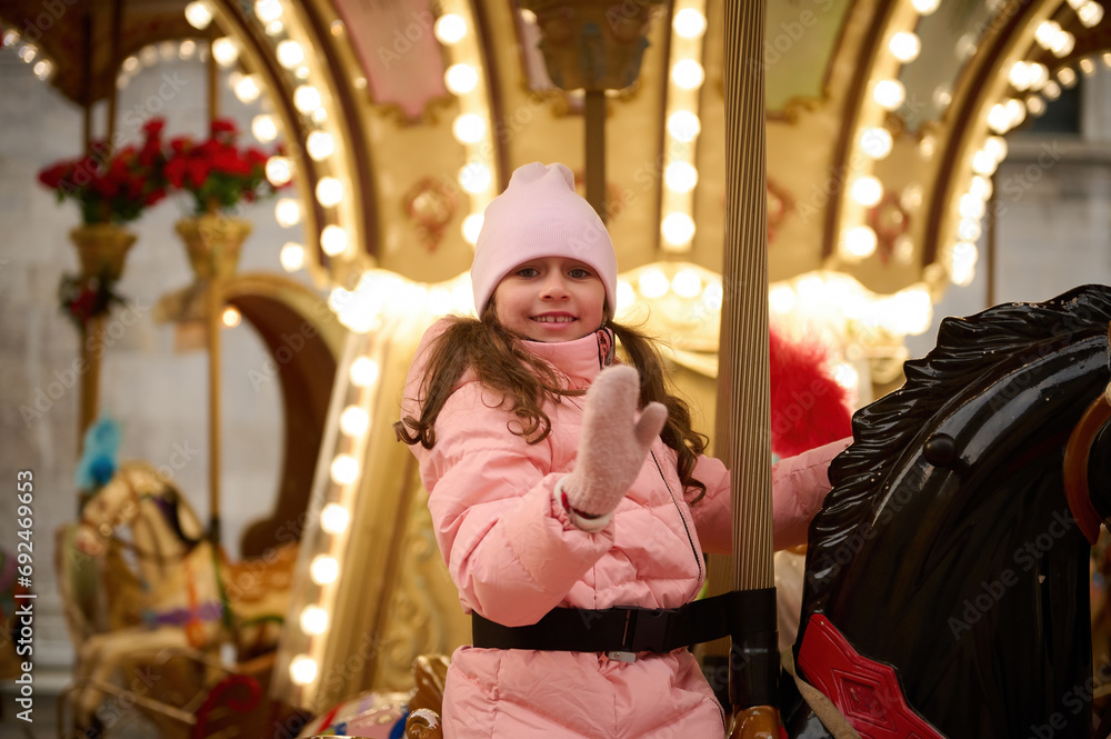 Cute smiling little child girl riding a wooden horse on merry go round carousel at a festive family Christmas market