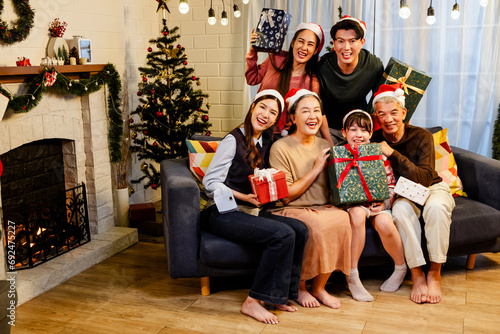Happy and cheerful Asian family gathering, wearing Santa hats talking and smiling. Exchanging gifts, grandparents, grandchildren, daughters, sitting at the fireplace with decorated Christmas tree.