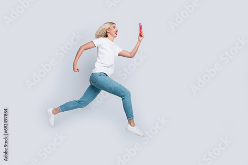 Full size photo of cheerful woman dressed white t-shirt denim pants fast run look at smartphone isolated on white color background