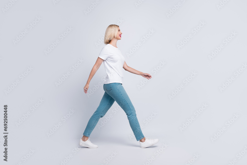 Full length photo of good mood gorgeous girl dressed white t-shirt denim pants walk look at empty space isolated on gray color background