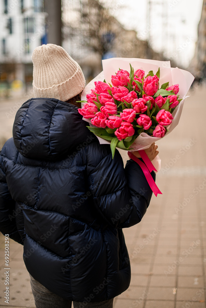 Fototapeta premium Tenderness pink tulips in the hands of a girl in a black jacket and a light beige hat
