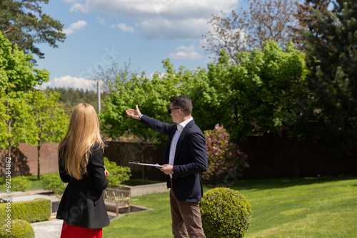 Real estate agent shows female client in formal suit lit cottage garden. Businesswoman listens to property manager house explanation