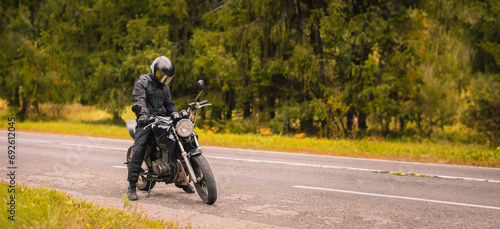 motorcyclist in motorcycle clothing and a helmet on a custom stylish motorcycle on a forest asphalt road