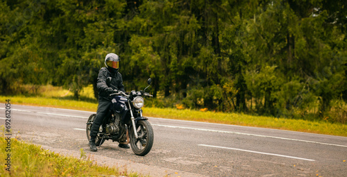 motorcyclist in motorcycle clothing and a helmet on a custom stylish motorcycle on a forest asphalt road