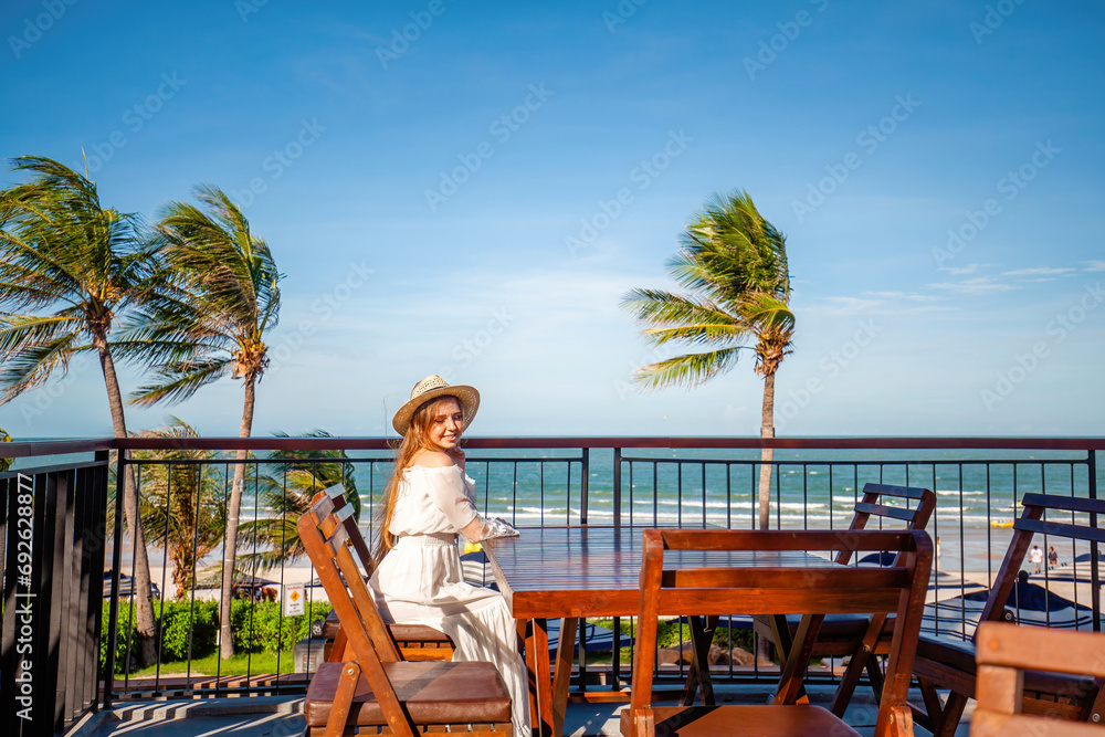 Woman in white dress with straw hat enjoying coastal view from wooden balcony. Tropical beach backdrop with swaying palm trees. Luxury travel destinations.