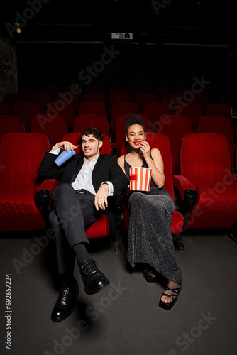 happy diverse couple in chic outfits enjoying movie and holding soda and popcorn while on date