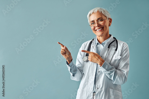 Beautiful confident mature gray-haired woman doctor in white medical coat and glasses standing on light blue background and pointing with fingers to free space in background and smiling.