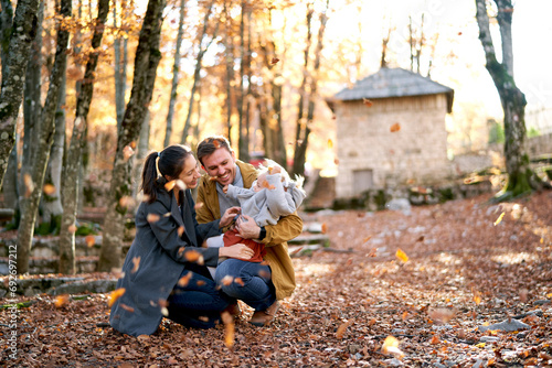 Mom crouches and hugs dad with a little girl on his knees in the park under falling leaves photo