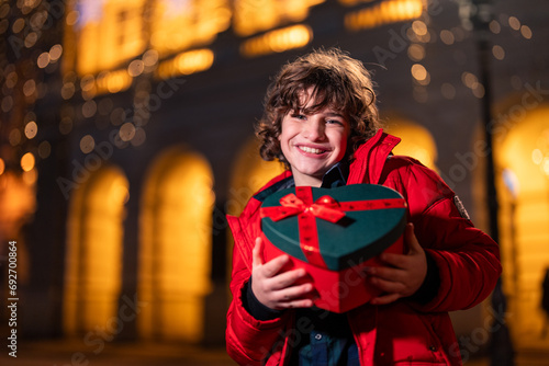 Smiling young boy holding Christmas gift box while standing outdoors. Excited young boy holding heart shaped gift box with ribbon standing in the city center.