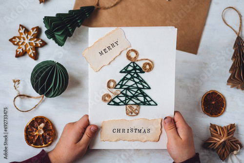 Concept of Christmas craft, handmade presents, family leisure time, hobby, recreation. Little kid with paper postcard with new year fir tree embroidering. Gingerbread cookies, lights, cozy atmosphere